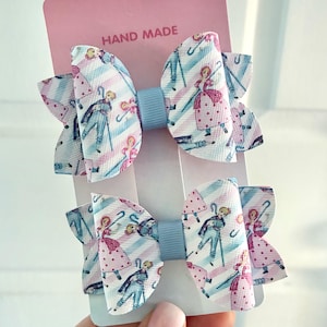 May include: Two hair bows attached to a pink display card labelled HAND MADE. The bows feature a repeating pattern of cartoon characters with blue canes and pink polka-dot dresses against diagonal light blue and white stripes, with blue centres.