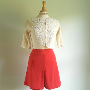 May include: A cream-colored cardigan with white floral embroidery, paired with a bright red A-line skirt with a button-up front.