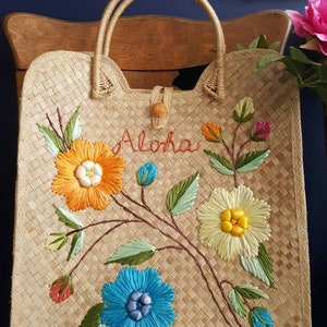 Puede incluir: Un bolso de mano de paja trenzada con flores bordadas y las palabras "Aloha Hawaii" en hilo rojo.