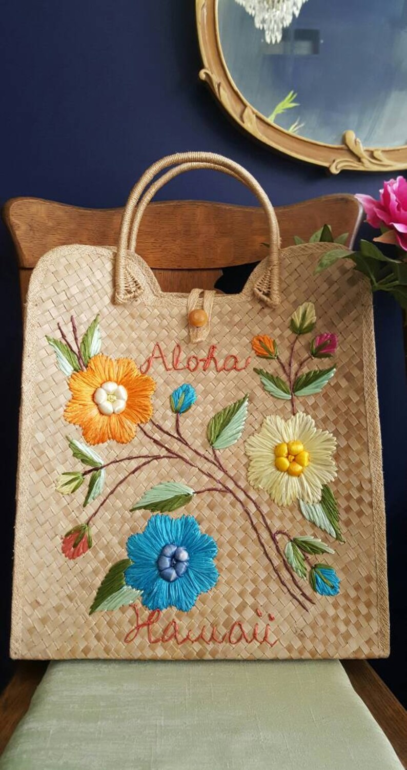 Puede incluir: Un bolso de mano de paja trenzada con flores bordadas y las palabras "Aloha Hawaii" en hilo rojo.