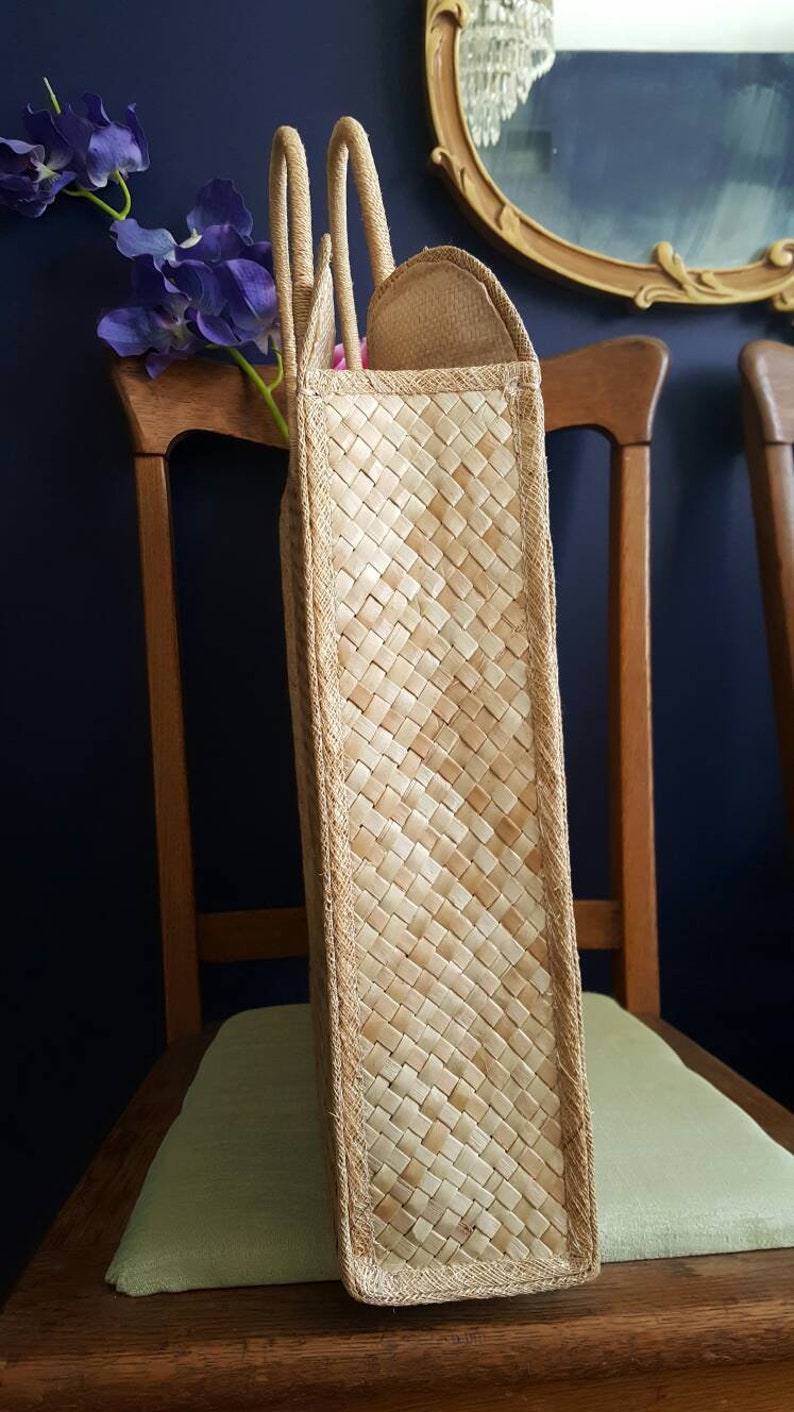 Puede incluir: Un bolso de mano de paja tejido con dos asas. El bolso es de color marr&oacute;n claro y tiene un patr&oacute;n tejido. El bolso est&aacute; sentado en una silla de madera.