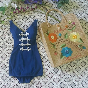 Puede incluir: Un traje de ba&ntilde;o azul con detalles de cuerda blanca y un escote en V. El traje de ba&ntilde;o est&aacute; sobre una superficie de crochet blanco junto a una bolsa de paja con flores bordadas de colores y el texto "Aloha Hawaii".