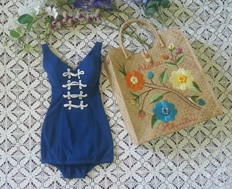 Puede incluir: Un traje de ba&ntilde;o azul con detalles de cuerda blanca y un escote en V. El traje de ba&ntilde;o est&aacute; sobre una superficie de crochet blanco junto a una bolsa de paja con flores bordadas de colores y el texto "Aloha Hawaii".