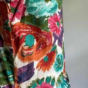 May include: A sleeveless garment featuring a vibrant floral pattern. The design showcases large, stylized flowers in shades of orange, pink, teal, and purple against a cream background. The fabric appears to be cotton or a similar material.