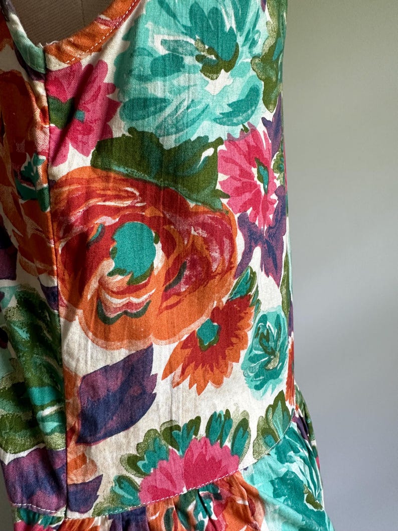 May include: A sleeveless garment featuring a vibrant floral pattern. The design showcases large, stylized flowers in shades of orange, pink, teal, and purple against a cream background. The fabric appears to be cotton or a similar material.