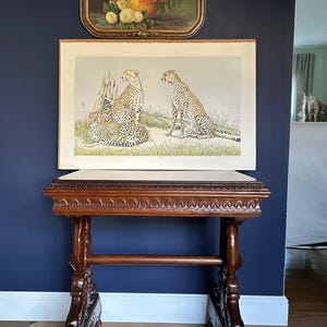 Puede incluir: Una mesa auxiliar de madera antigua con tallas ornamentadas, coronada por una impresión enmarcada de guepardos. Arriba, una naturaleza muerta floral enmarcada cuelga contra una pared azul oscuro. La mesa tiene un acabado marrón oscuro.