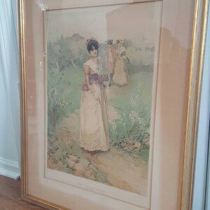Puede incluir: Una impresi&oacute;n enmarcada de una mujer con un vestido blanco caminando por un jard&iacute;n. La impresi&oacute;n est&aacute; en un marco dorado y tiene un passe-partout blanco. El t&iacute;tulo de la impresi&oacute;n es "The Garden Gate".