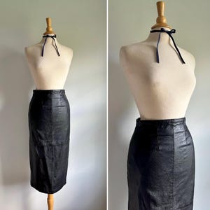May include: A black leather pencil skirt displayed on a mannequin. The skirt is knee-length and has a fitted silhouette. A dark blue ribbon is tied around the mannequin's neck. The background is a plain, light-colored wall.