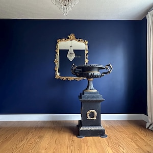 Puede incluir: Una urna decorativa negra y dorada con una base de pedestal se encuentra en un suelo de madera dura frente a una pared azul oscuro. Un espejo dorado enmarcado con un reflejo de araña de cristal cuelga de la pared.