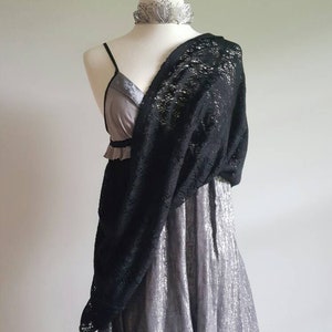 K&ouml;nnte beinhalten: Ein silbernes Kleid mit einem schwarzen Spitzen-Schal, der dar&uuml;ber gelegt ist. Das Kleid hat einen R&uuml;schen-Ausschnitt und ein tailliertes Oberteil. Der Schal ist aus zarter Spitze und hat ein flie&szlig;endes Design.