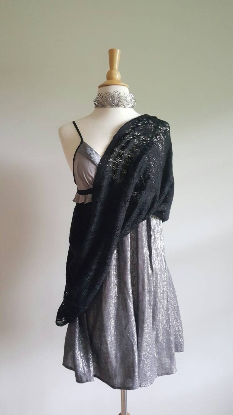 K&ouml;nnte beinhalten: Ein silbernes Kleid mit einem schwarzen Spitzen-Schal, der dar&uuml;ber gelegt ist. Das Kleid hat einen R&uuml;schen-Ausschnitt und ein tailliertes Oberteil. Der Schal ist aus zarter Spitze und hat ein flie&szlig;endes Design.