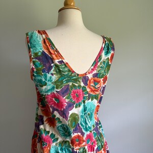 May include: A sleeveless floral dress with a V-neckline, displayed on a mannequin. The dress features a vibrant pattern of large flowers in shades of pink, orange, teal, and purple against a white background. The dress has a pleated skirt.