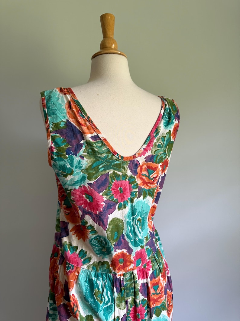 May include: A sleeveless floral dress with a V-neckline, displayed on a mannequin. The dress features a vibrant pattern of large flowers in shades of pink, orange, teal, and purple against a white background. The dress has a pleated skirt.