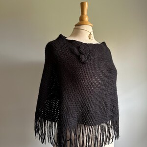 May include: Black knitted poncho with a fringe trim and a flower detail on the neckline.
