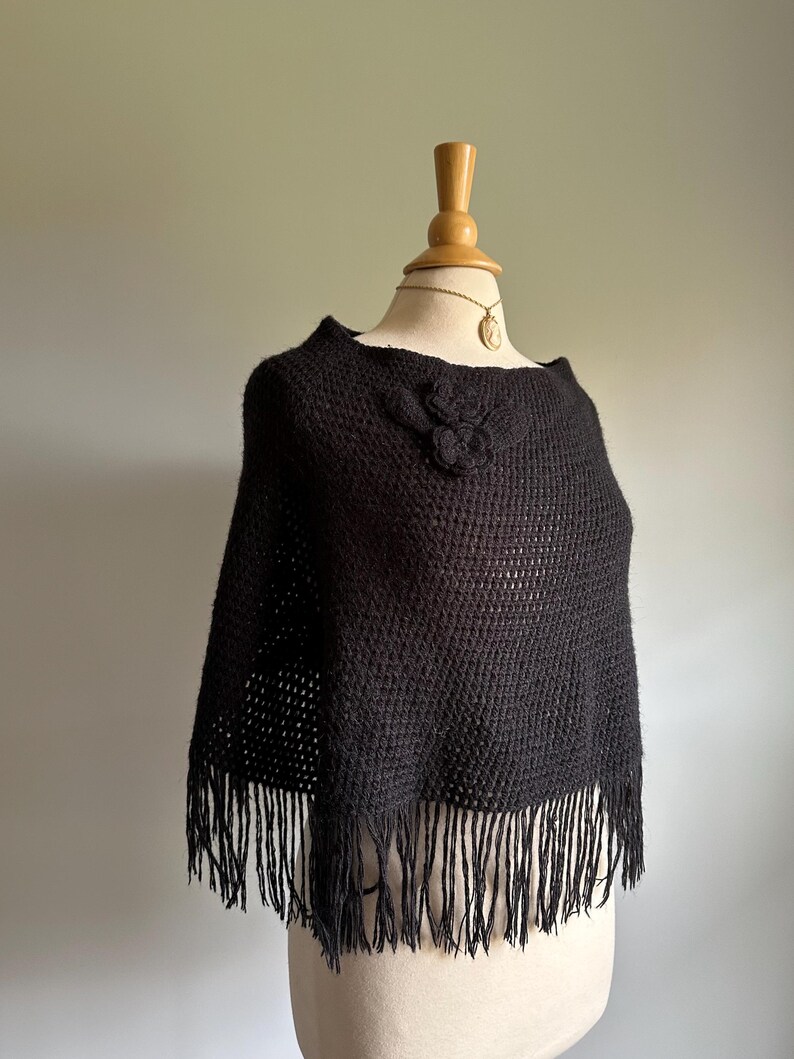 May include: Black knitted poncho with a fringe trim and a flower detail on the neckline.