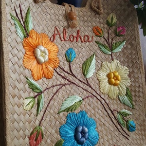 Puede incluir: Un bolso de mano de paja trenzada con flores bordadas y las palabras "Aloha Hawaii". Las flores son de color naranja, amarillo, azul y verde.