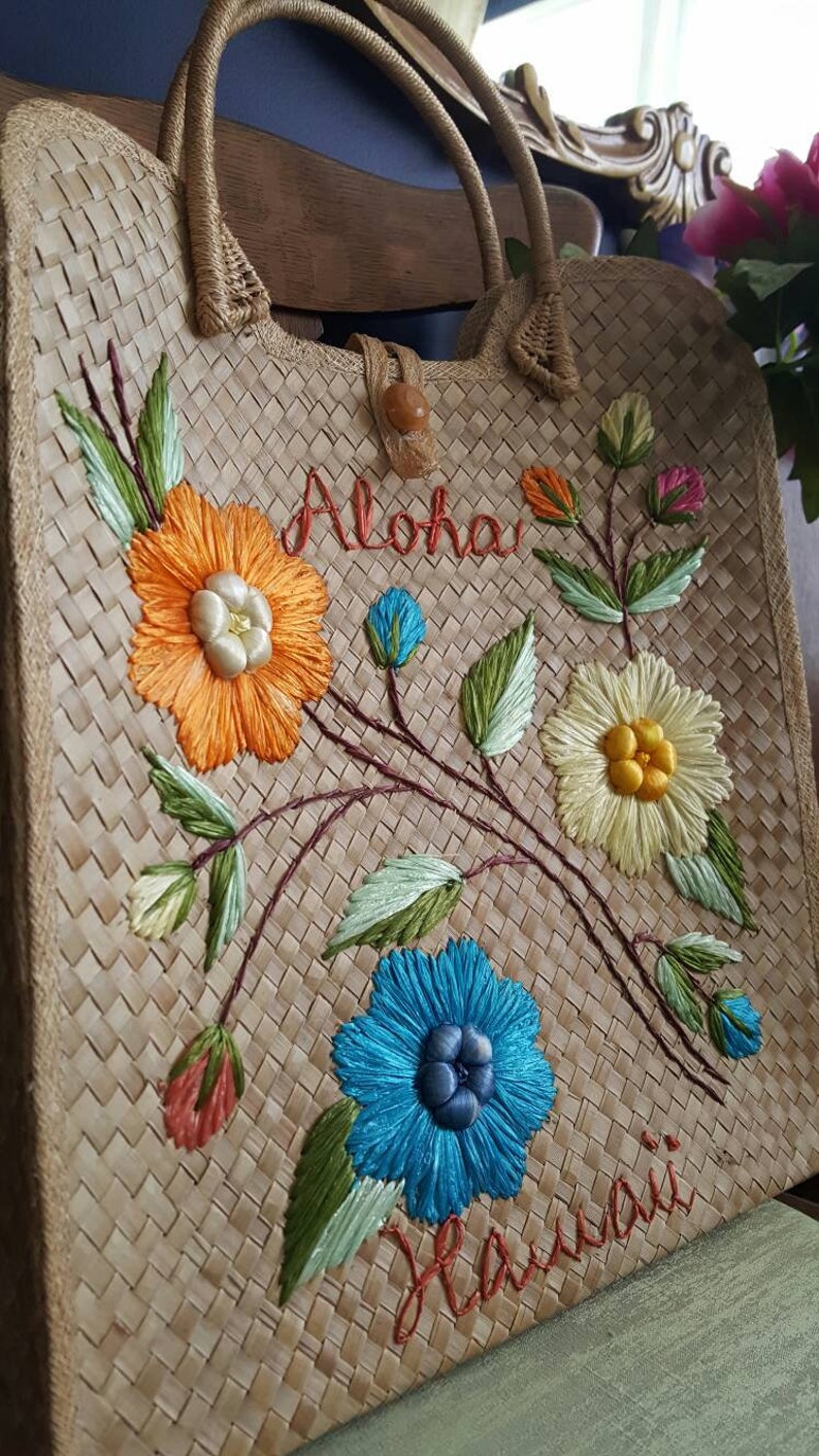Puede incluir: Un bolso de mano de paja trenzada con flores bordadas y las palabras "Aloha Hawaii". Las flores son de color naranja, amarillo, azul y verde.