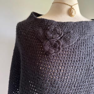 May include: Black crochet poncho with a large black flower embellishment. The poncho has a loose fit and a wide neckline.