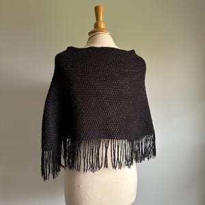 May include: Black knitted poncho with fringe trim. The poncho is displayed on a white mannequin.