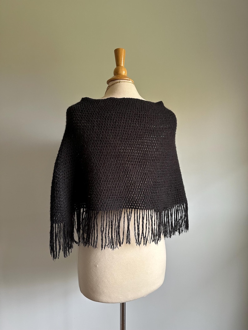 May include: Black knitted poncho with fringe trim. The poncho is displayed on a white mannequin.