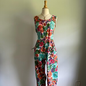 May include: A sleeveless floral jumpsuit in a vibrant mix of colors, including orange, teal, purple, and pink. The jumpsuit features a button-down front, pockets, and a fitted waist. The floral pattern covers the entire garment.
