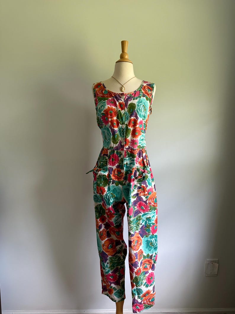 May include: A sleeveless floral jumpsuit in a vibrant mix of colors, including orange, teal, purple, and pink. The jumpsuit features a button-down front, pockets, and a fitted waist. The floral pattern covers the entire garment.
