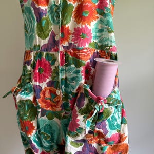 May include: A sleeveless floral jumpsuit with a vibrant pattern of orange, pink, teal, and purple flowers on a white background. The jumpsuit has pockets with tie details and a button-up front. A roll of pink tulle is in the pocket.