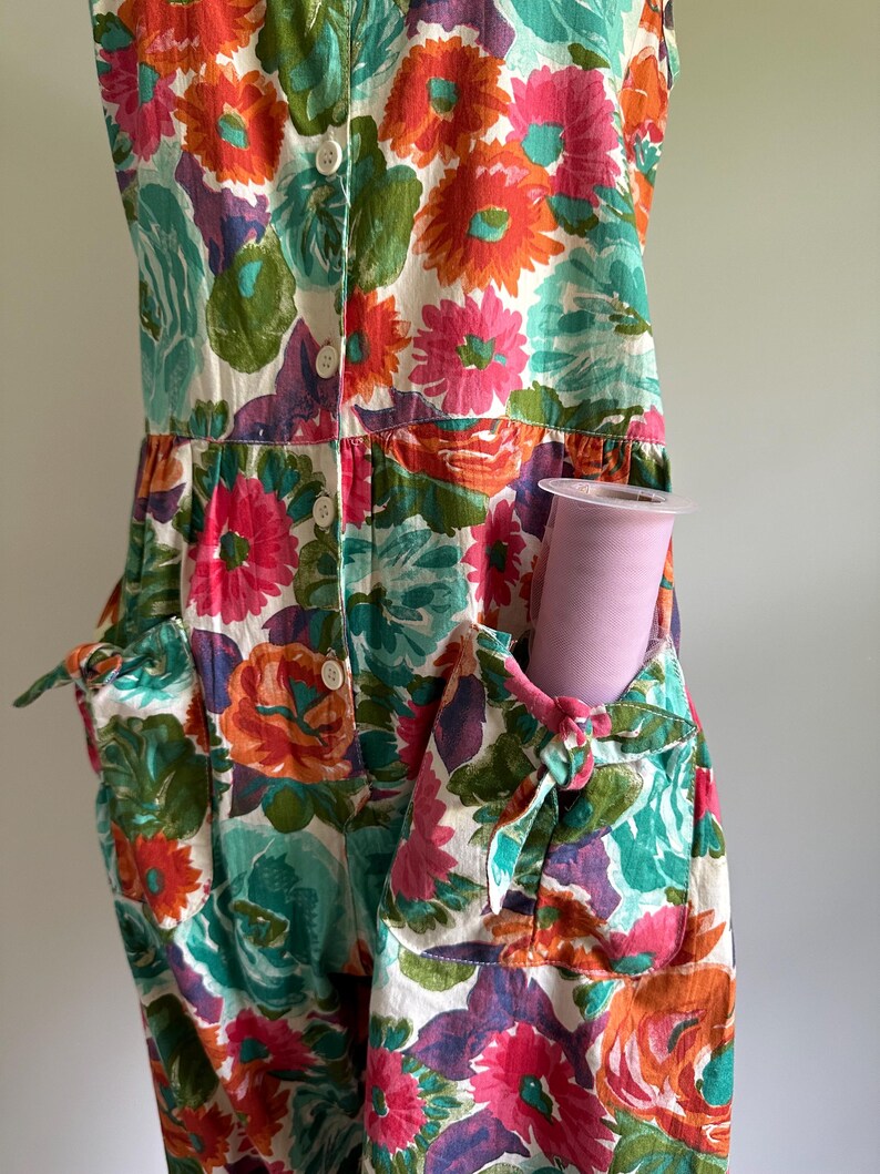 May include: A sleeveless floral jumpsuit with a vibrant pattern of orange, pink, teal, and purple flowers on a white background. The jumpsuit has pockets with tie details and a button-up front. A roll of pink tulle is in the pocket.