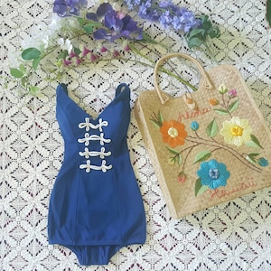 Op de afbeelding: Een blauwe badpak met witte touwdetails en een diepe V-hals. Het badpak ligt op een wit gehaakt tafelkleed met een strooien tas geborduurd met bloemen en de woorden "Aloha Hawaii".