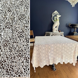 Puede incluir: Un mantel de ganchillo color crema con un estampado floral. El intrincado diseño de encaje cubre una gran mesa cuadrada, con los bordes de la tela colgando. El mantel está sobre un suelo de madera.