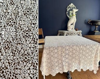 Könnte beinhalten: Eine cremefarbene, gehäkelte Tischdecke mit floralem Muster. Das filigrane Spitzendesign bedeckt einen großen quadratischen Tisch, wobei die Kanten des Stoffes herunterhängen. Die Tischdecke liegt auf einem Holzboden.
