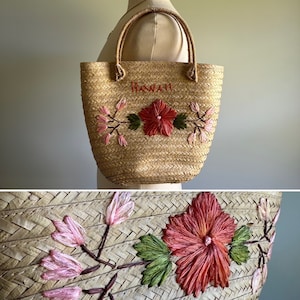 Op de afbeelding: Een strooien tas met geborduurde roze en rode bloemen en groene bladeren. De tas heeft het woord "Hawaii" geborduurd op de voorkant.