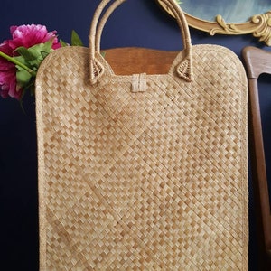 Puede incluir: Un bolso de mano de paja trenzada de color marr&oacute;n claro con dos asas. El bolso est&aacute; sobre una superficie de madera con un coj&iacute;n verde.