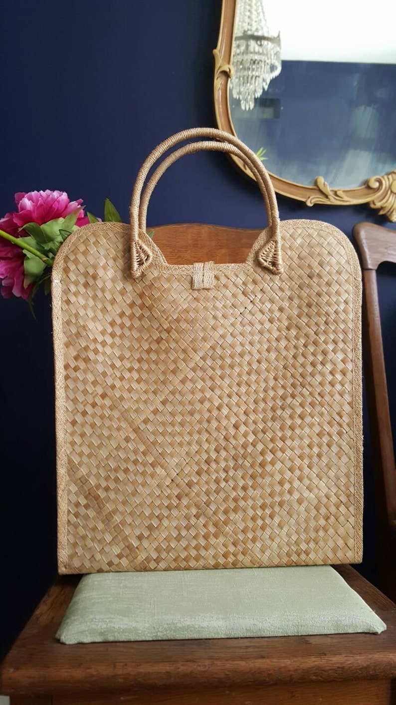 Puede incluir: Un bolso de mano de paja trenzada de color marr&oacute;n claro con dos asas. El bolso est&aacute; sobre una superficie de madera con un coj&iacute;n verde.