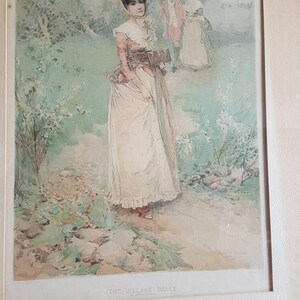 Puede incluir: Una impresi&oacute;n enmarcada de una pintura de acuarela que representa a una mujer con un vestido blanco y un sombrero rojo caminando por un jard&iacute;n. La pintura se titula "The Village Belle" y est&aacute; firmada por el artista.