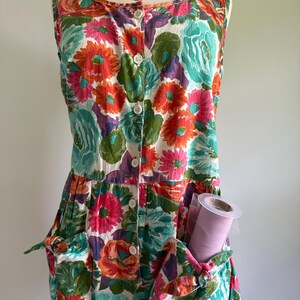 May include: A sleeveless floral print dress with a button-down front and two front pockets. The dress features a vibrant pattern of orange, pink, teal, and purple flowers on a white background. A roll of pink tulle is visible in one of the pockets.