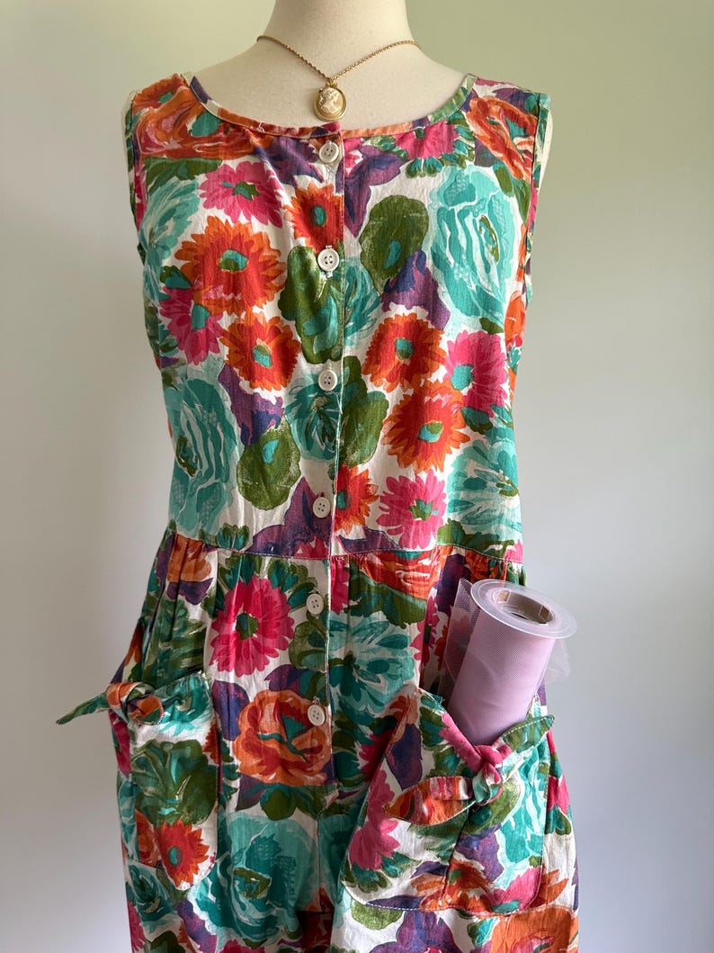 May include: A sleeveless floral print dress with a button-down front and two front pockets. The dress features a vibrant pattern of orange, pink, teal, and purple flowers on a white background. A roll of pink tulle is visible in one of the pockets.