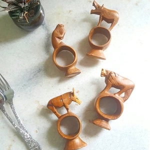 Puede incluir: Conjunto de cuatro anillos de servilleta de madera, cada uno con un animal tallado diferente: un mono, un jabalí, un león y una cebra. Los anillos están hechos de madera marrón oscura y tienen un aspecto rústico y artesanal.