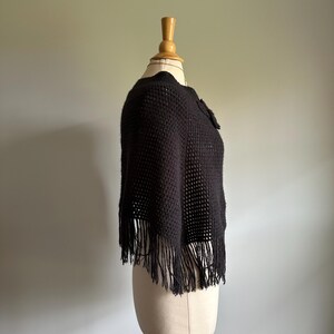 May include: A black crocheted poncho with fringe on a mannequin. The poncho has a loose fit and a simple design.