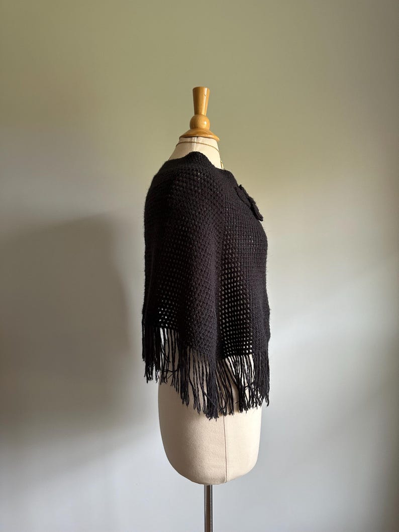 May include: A black crocheted poncho with fringe on a mannequin. The poncho has a loose fit and a simple design.