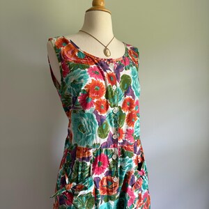 May include: A sleeveless floral print dress with a button-down front and pockets. The dress features a vibrant pattern of orange, pink, teal, and purple flowers on a white background. A gold-toned pendant necklace is visible at the neckline. The dress is displayed on a mannequin.