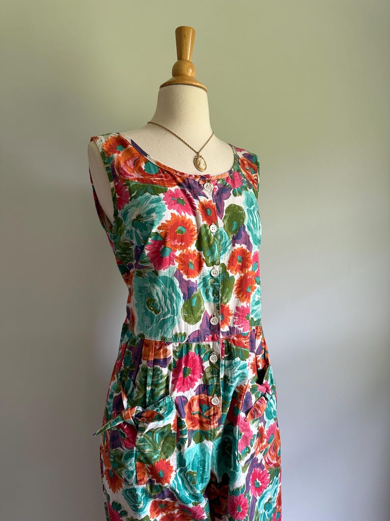 May include: A sleeveless floral print dress with a button-down front and pockets. The dress features a vibrant pattern of orange, pink, teal, and purple flowers on a white background. A gold-toned pendant necklace is visible at the neckline. The dress is displayed on a mannequin.