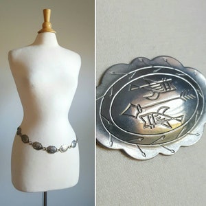 以下が含まれることがあります： 矢印と鳥のスタイリッシュなデザインが施された、楕円形のリンクを持つシルバーチェーンベルト。ベルトは白いマネキンに飾られています。