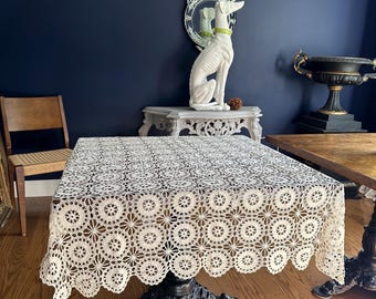 Könnte beinhalten: Eine cremefarbene, gehäkelte Spitzendecke bedeckt einen dunklen, verzierten Tisch. Die Tischdecke hat einen gewellten Rand und ein sich wiederholendes Blumenmuster. Der Tisch hat ein schwarzes Gusseisen-Gestell. Der Raum hat Holzböden und eine dunkelblaue Wand.