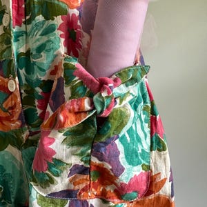 May include: A floral print dress with a pocket holding a roll of pink tulle. The dress features a vibrant pattern of pink, orange, green, and purple flowers on a white background. The pocket is tied with a pink knot.