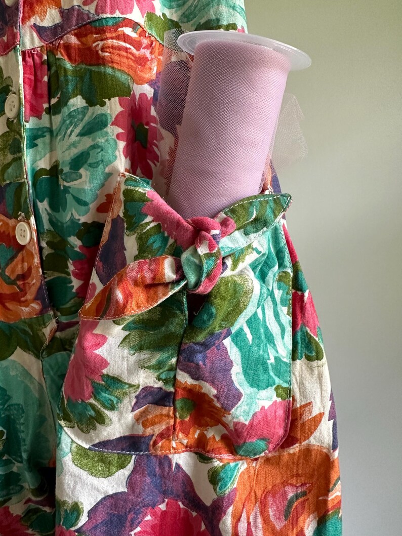 May include: A floral print dress with a pocket holding a roll of pink tulle. The dress features a vibrant pattern of pink, orange, green, and purple flowers on a white background. The pocket is tied with a pink knot.