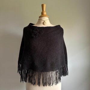 May include: Black knitted poncho with a flower detail and fringe trim.