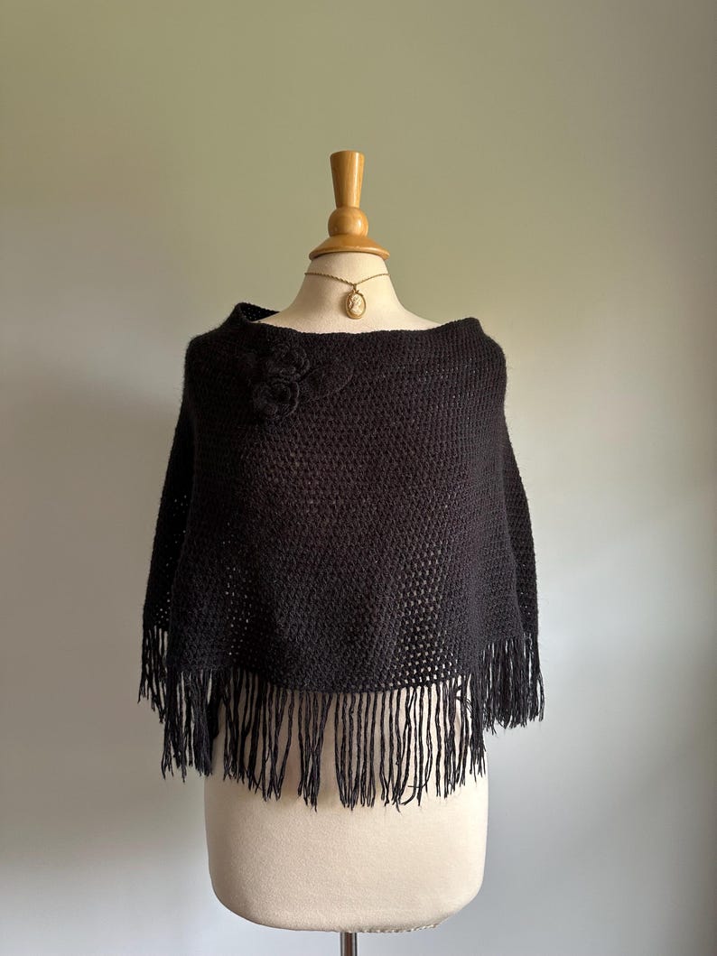 May include: Black knitted poncho with a flower detail and fringe trim.