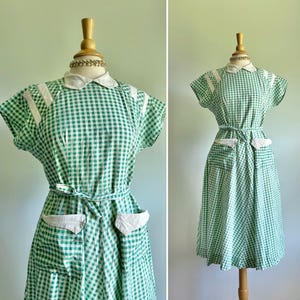 May include: A vintage green and white gingham dress displayed on a mannequin. The dress features a white collar, white shoulder accents, a matching belt, and two white pocket details. The dress has short sleeves and a knee-length skirt.