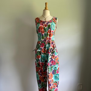 May include: A sleeveless floral jumpsuit in a vibrant pattern of red, orange, teal, and purple flowers on a white background. The jumpsuit features a button-down front, pockets, and a fitted waist. The garment is displayed on a mannequin.
